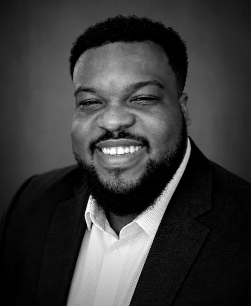 Zachary Stovall smiling in a dark suit jacket and light shirt, in black and white.
